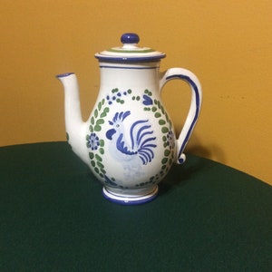May include: White ceramic teapot with a blue and green rooster design. The teapot has a curved handle, a spout, and a lid with a blue knob. The rooster is surrounded by green leaves and blue flowers.