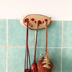 Puede incluir: Un colgante de pared de madera con un acabado de vetas naturales, adornado con detalles de flores de amapola rojas. Tres tallos decorativos cuelgan debajo, cada uno con elementos florales secos &uacute;nicos, incluyendo c&aacute;psulas de semillas y follaje texturizado. El fondo es una pared de azulejos azul claro.