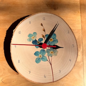 May include: A round wooden clock with a light wood grain. The clock face features blue floral accents and black numerals. The clock hands are black and red, with a red center. The clock is approximately 8 inches in diameter.