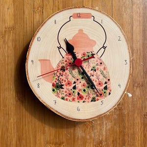 Può includere: Un orologio in legno con un design a forma di teiera con motivo floreale. Il quadrante dell'orologio ha numeri neri e lancette nere. La teiera è rosa con fiori verdi e rosa.