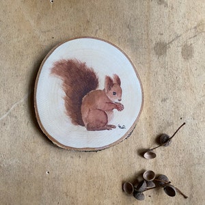 Puede incluir: Una pintura de acuarela de una ardilla marrón sobre una rodaja de madera redonda. La ardilla está mirando hacia la derecha y tiene una cola tupida. El fondo es de madera marrón claro.