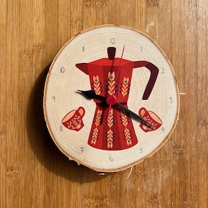 Op de afbeelding: Een houten klok met een rood en wit koffiekanontwerp. De wijzerplaat van de klok heeft zwarte cijfers en wijzers. Twee kleine kopjes koffie bevinden zich aan weerszijden van de koffiekan.