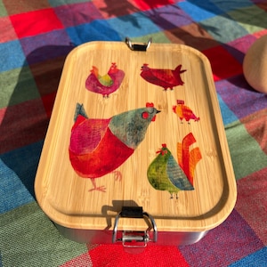 Può includere: Un portapranzo rettangolare con coperchio in bambù con illustrazioni colorate di galline. Il coperchio è fissato con un fermaglio in metallo. Il portapranzo è in metallo e bambù.