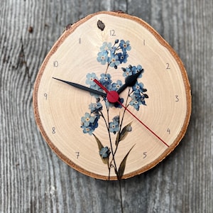 Può includere: Un orologio rotondo in legno con finitura a venatura naturale. Il quadrante presenta un disegno floreale di nontiscordardimé blu e lancette nere con una lancetta dei secondi rossa. I numeri sono stampati in nero.