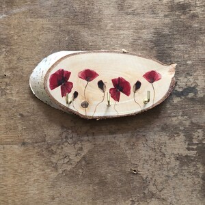 Puede incluir: Un llavero de madera con borde de corteza natural. Presenta cinco dise&ntilde;os de flores de amapola roja y ganchos de lat&oacute;n para llaves. La madera tiene un acabado claro y las amapolas tienen un efecto acuarela. Mide aproximadamente 20 cm de largo.