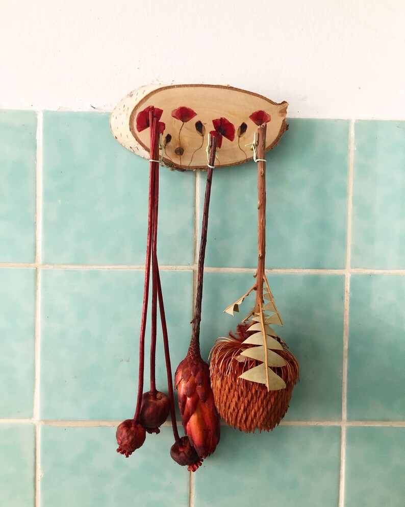 Puede incluir: Un colgante de pared de madera con un acabado de vetas naturales, adornado con detalles de flores de amapola rojas. Tres tallos decorativos cuelgan debajo, cada uno con elementos florales secos &uacute;nicos, incluyendo c&aacute;psulas de semillas y follaje texturizado. El fondo es una pared de azulejos azul claro.