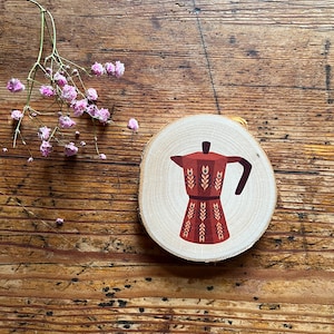 Könnte beinhalten: Ein Holz-Untersetzer mit einer roten und weißen Illustration einer Kaffeekanne mit floralem Muster. Der Untersetzer ist rund und hat eine natürliche Holzoptik.