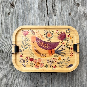 Op de afbeelding: Een rechthoekige bamboe lunchbox met een vogel- en bloemenontwerp. De vogel is oranje en paars, omringd door roze, rode en gele bloemen. De doos heeft zwarte metalen sluitingen.