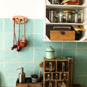Puede incluir: Una escena de cocina con un especiero de madera con recipientes de vidrio, un peque&ntilde;o cubo de metal y un dispensador de jab&oacute;n. Una estanter&iacute;a de madera contiene tarros de t&eacute; y especias. El fondo tiene azulejos turquesa y una encimera de madera.