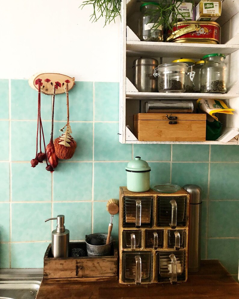 Puede incluir: Una escena de cocina con un especiero de madera con recipientes de vidrio, un peque&ntilde;o cubo de metal y un dispensador de jab&oacute;n. Una estanter&iacute;a de madera contiene tarros de t&eacute; y especias. El fondo tiene azulejos turquesa y una encimera de madera.