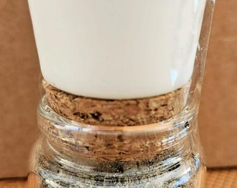 May include: A small glass jar filled with dried herbs, a cork stopper, and a white ceramic cup on top. The jar has a label that reads "Baneful Herbs, Rose Quartz, Black Agate. Hex or Curse."