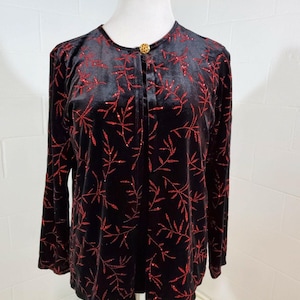 May include: Black velvet long-sleeve top with a red leaf pattern. The top has a round neckline and a single gold button closure. The red leaf design is glittery, adding a touch of sparkle to the garment.