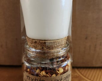 May include: A small glass jar filled with red flakes and a cork stopper, topped with a white candle. The jar has a handwritten label with the words "Tektite black pepper" and other ingredients. The candle is wrapped in clear plastic.