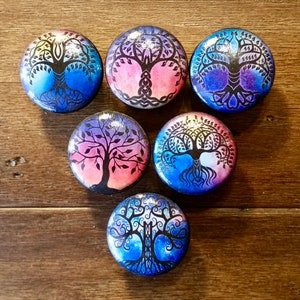 May include: Six round wooden coasters with a watercolor tree of life design. Each coaster features a different color scheme with a black silhouette of the tree. The coasters are perfect for adding a touch of nature to your home decor.