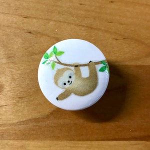 May include: A white round button with a cartoon sloth hanging from a branch with green leaves. The sloth is brown with a white face and a smile.