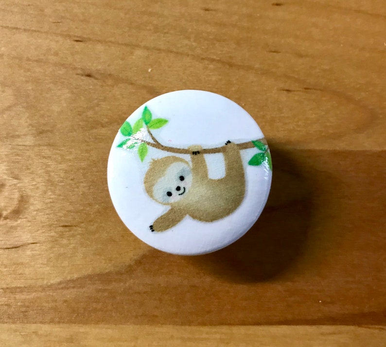 May include: A white round button with a cartoon sloth hanging from a branch with green leaves. The sloth is brown with a white face and a smile.