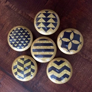 May include: Six decorative wooden cabinet knobs with gold rims and various geometric patterns in black and gold. Designs include triangles, stripes, and chevrons. The knobs are arranged on a dark wood surface.