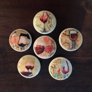 May include: Six round ceramic knobs with vintage-inspired designs featuring wine glasses and grapes. The knobs have a white background with a distressed look and are decorated with red, brown, and gold accents.