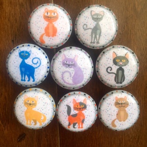 May include: Nine round ceramic knobs with a white background and colorful cat illustrations. Each knob has a white polka dot pattern and a black border.