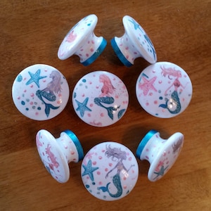 May include: Set of eight white ceramic cabinet knobs with a mermaid design. Each knob features a mermaid with a pink tail and a blue top, surrounded by pink and blue dots, and a starfish. The knobs have a turquoise trim.