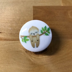 May include: A white round button with a cartoon illustration of a brown sloth hanging upside down from a branch with green leaves.