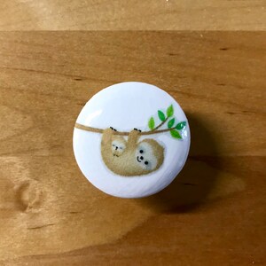 May include: A white round button with a cartoon image of a mother sloth and her baby sloth hanging upside down from a branch with green leaves.