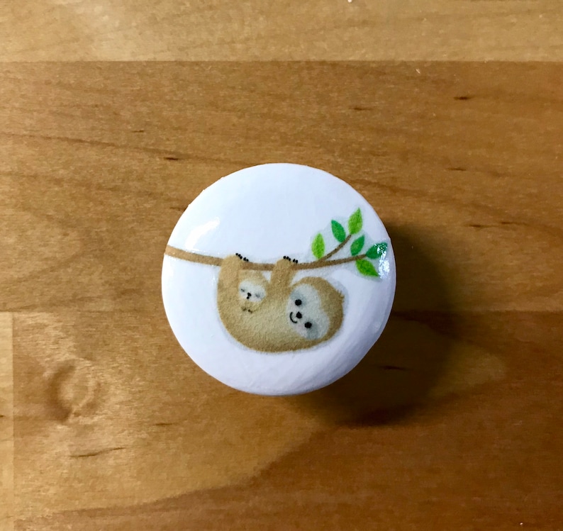 May include: A white round button with a cartoon image of a mother sloth and her baby sloth hanging upside down from a branch with green leaves.