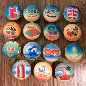 May include: Twelve round wooden knobs with various summer-themed designs. The designs include a swimsuit, flip flops, a beach umbrella, a wave, a life preserver, an anchor, a beach bar, a beach hut, sunglasses, and text that says "Summer", "Beach", and "Recreation Time".