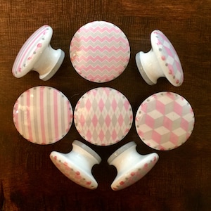 May include: Set of eight pink and gray patterned cabinet knobs. The knobs have a white base and are decorated with various geometric patterns, including stripes, chevrons, diamonds, and cubes.
