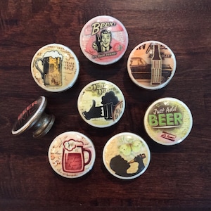May include: Nine ceramic cabinet knobs with various beer-themed designs. The knobs are round and feature images of beer bottles, glasses, and other beer-related imagery. The knobs are in various colors, including brown, white, and green.