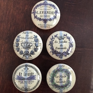 May include: Four round, white, decorative buttons with lavender floral designs and French text. The buttons feature the words "Confiserie des Tuileries Lavande au Miel", "Savon de Marseille", "Le Jardin de Lavande", and "Le Jardin Le Fleur de Lavande".