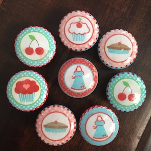 Puede incluir: Nueve pomos redondos de cerámica decorativos de varios colores con imágenes de cerezas, magdalenas, tartas y delantales. Los pomos tienen un fondo blanco con detalles rojos y azules.