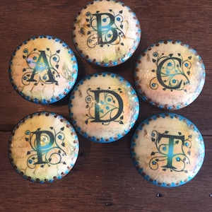 May include: Six round wooden knobs with a vintage design. Each knob features a different letter of the alphabet, A, B, C, D, P, and T, in a black script font with a floral design. The knobs are painted in a cream color with a brown border and a turquoise blue trim.