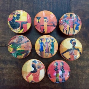 May include: Nine round wooden coasters with colorful African-inspired designs featuring women in various poses. The coasters have a brown background with yellow, orange, and red accents.