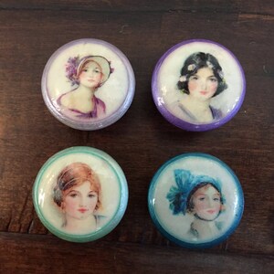 May include: Four round, decorative, painted ceramic tiles with images of women wearing hats. The tiles have a white background with a purple, blue, or green border.