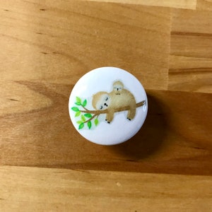 May include: A white button with a cartoon illustration of two sloths sleeping on a branch with green leaves.