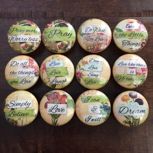 May include: A collection of twelve round decorative buttons with inspirational quotes. Each button features floral designs and phrases like "Pray," "Love," and "Dream" in various fonts and colors. The buttons are arranged on a dark wooden surface.