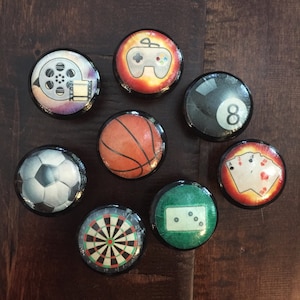 May include: A collection of eight round cabinet knobs with various designs. The knobs feature images of a film reel, a video game controller, an eight ball, a basketball, a soccer ball, playing cards, a dartboard, and dominoes. Each knob has a black border.