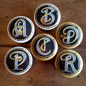 May include: Six round black and gold buttons with white polka dots around the edge. Each button features a white letter in a gold script font on a black background. The letters are A, B, C, D, P, and R.