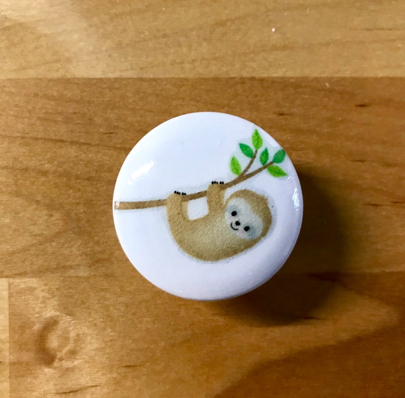 May include: A white ceramic button with a brown sloth hanging from a green branch. The sloth has a smiling face.