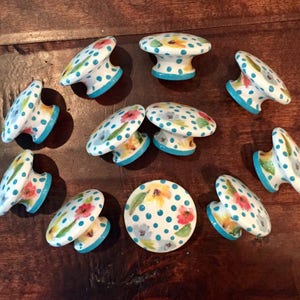 May include: Twelve white ceramic cabinet knobs with a turquoise trim. Each knob features a floral design with yellow, pink, and blue flowers and white polka dots.