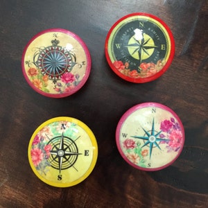 May include: Four round wooden coasters with compass designs and floral accents. Each coaster has a different colored border: red, pink, yellow, and black. The compass designs feature a black compass rose with a blue needle pointing north. The floral accents are in shades of pink, red, and yellow.