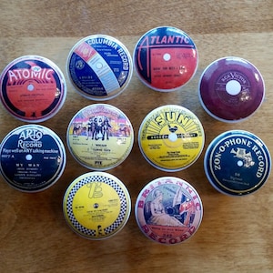 May include: Nine vintage record label designs printed on round white ceramic knobs. The labels feature various colors, including red, yellow, blue, and black, and include text such as "Columbia Record", "Atomic", "Arto Universal Record", "Atlantic", "Sun Record Company", "RCA Victor", "Zon-O-Phone Record", "Pye", and "Britannic Record".