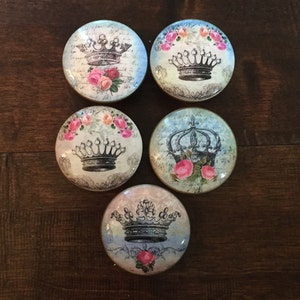 Puede incluir: Cinco pomos redondos decorativos para cajones con un diseño vintage. Cada pomo presenta una corona negra con rosas rosas y un fondo blanco. Los pomos tienen un borde de metal dorado.