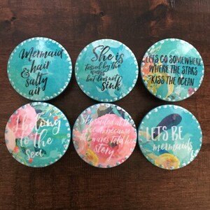 May include: Six round, turquoise blue, decorative coasters with white and pink floral designs and inspirational quotes. The quotes are: "Mermaid & hair salty air", "She is tossed by the waves but does not sink", "Let's go somewhere where the stars kiss the ocean", "Belong to the sea", "She smiled at the ocean because the waves told her story", and "Let's be mermaids".