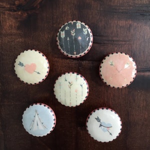 May include: Six round ceramic coasters with pink and black polka dot borders. Each coaster features a different design with arrows, hearts, and a teepee.