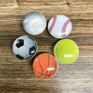 May include: Five different sports balls, each with a different design. The balls are arranged in a circle on a wooden surface. The balls are a volleyball, baseball, soccer ball, tennis ball, and basketball.