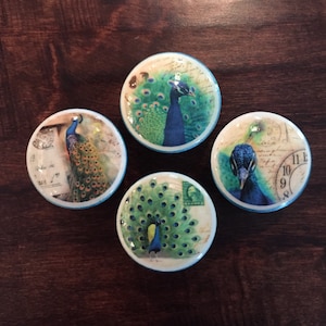 May include: Four round ceramic knobs with a blue and green peacock design. Each knob features a different peacock image, including a peacock with its tail feathers spread, a peacock with its head turned, and a peacock with its tail feathers partially spread. The knobs have a light blue trim around the edges.