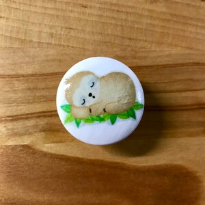 May include: A white round button with a cartoon illustration of a brown sloth sleeping on green leaves.