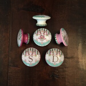 May include: Six decorative owl themed cabinet knobs with pink, white, and green accents. The knobs feature a variety of owl designs and the letters "S" and "D".